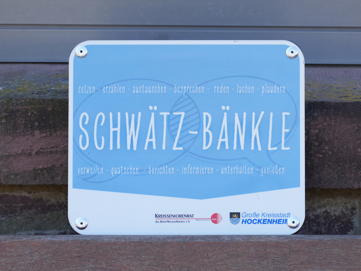 Blaues Schild mit der Aufschrift „Schwätz-Bänkle“ an einer Sitzbank in Hockenheim.